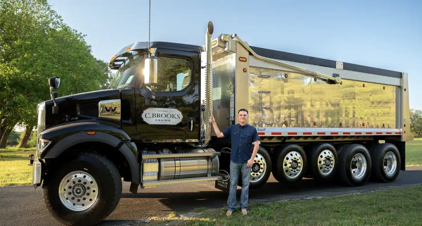 Man standing beside a shiny black-and-silver dump truck in a sunny outdoor setting