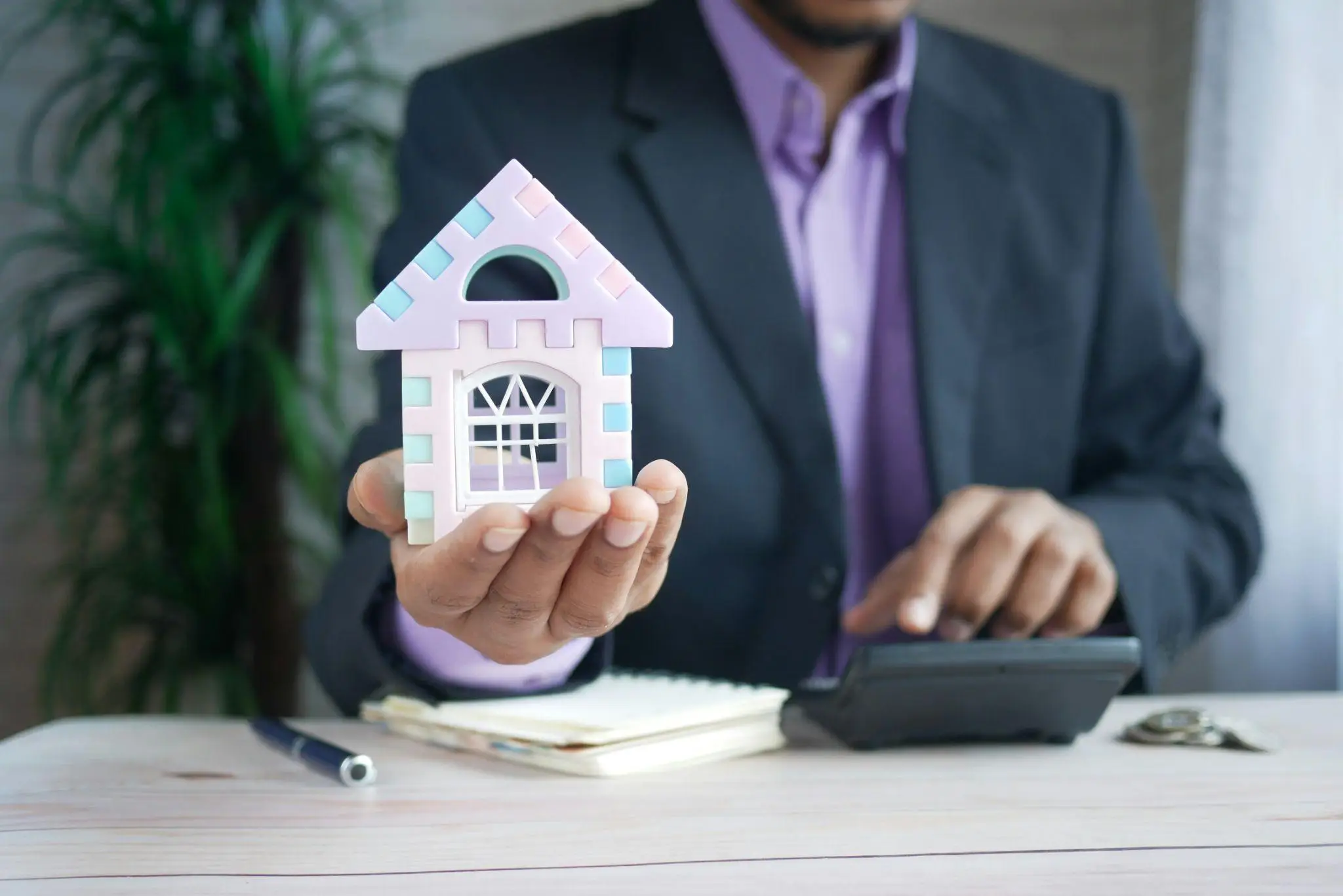 Small colorful toy house held by man in suit using calculator at desk