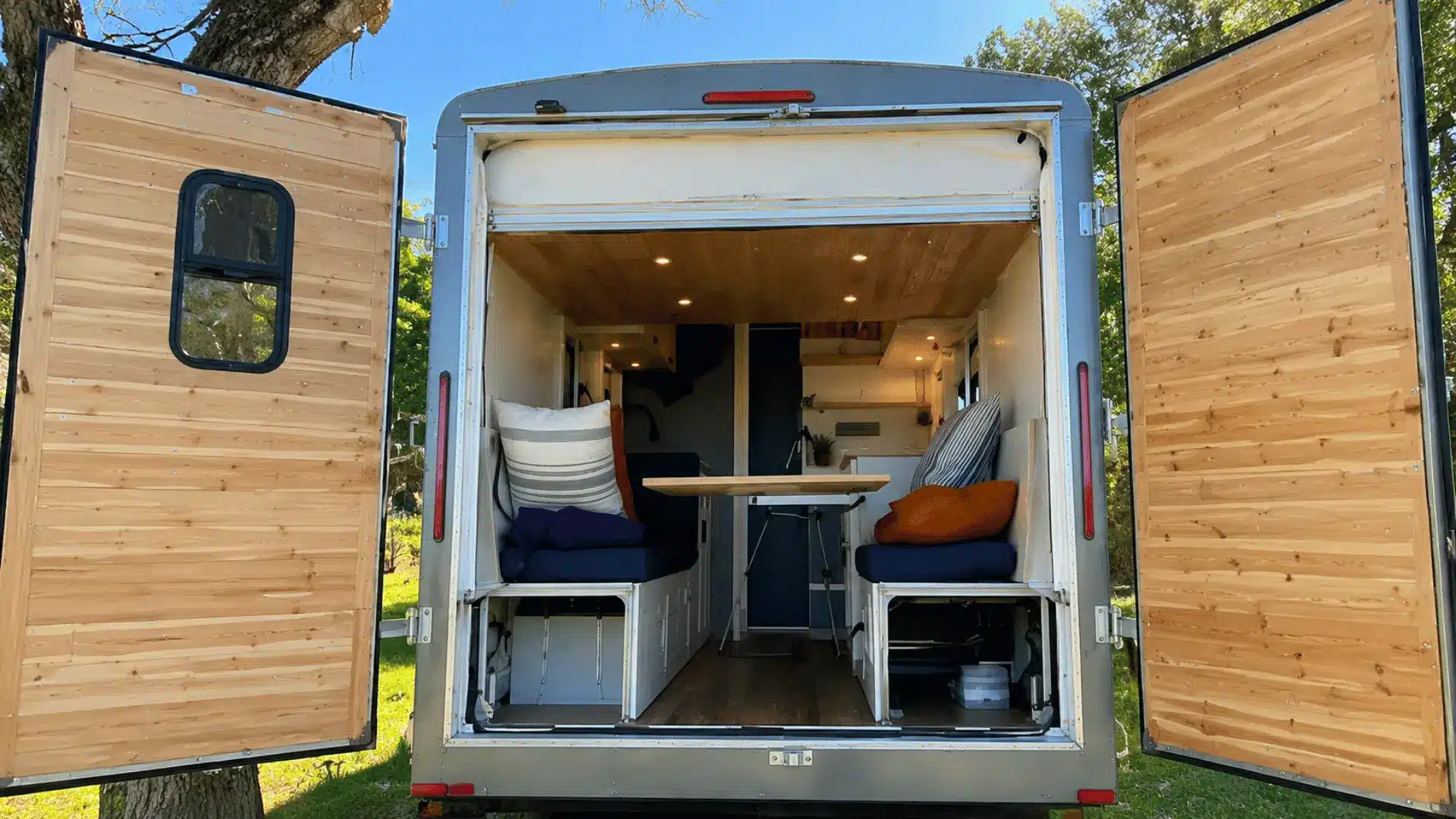 Cargo trailer camper interior with open rear doors, seating areas, table, and storage space.