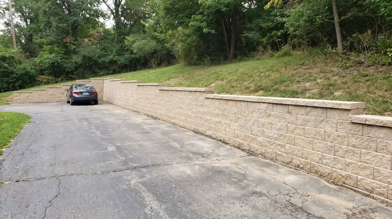 Concrete block retaining wall beside an asphalt driveway with a raised grassy slope and wooded background
