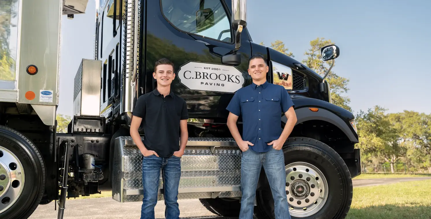 Two men standing in front of a black paving truck outdoors on a sunny day