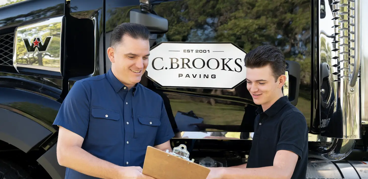 Two men discussing with clipboard beside black truck with paving company logo
