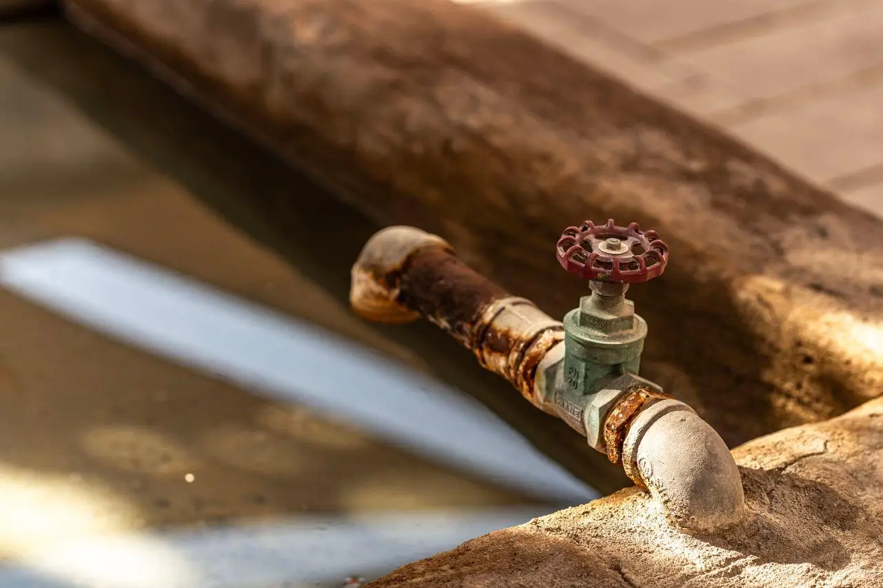 Rusty water pipe with red valve over shallow stone basin in warm sunlight