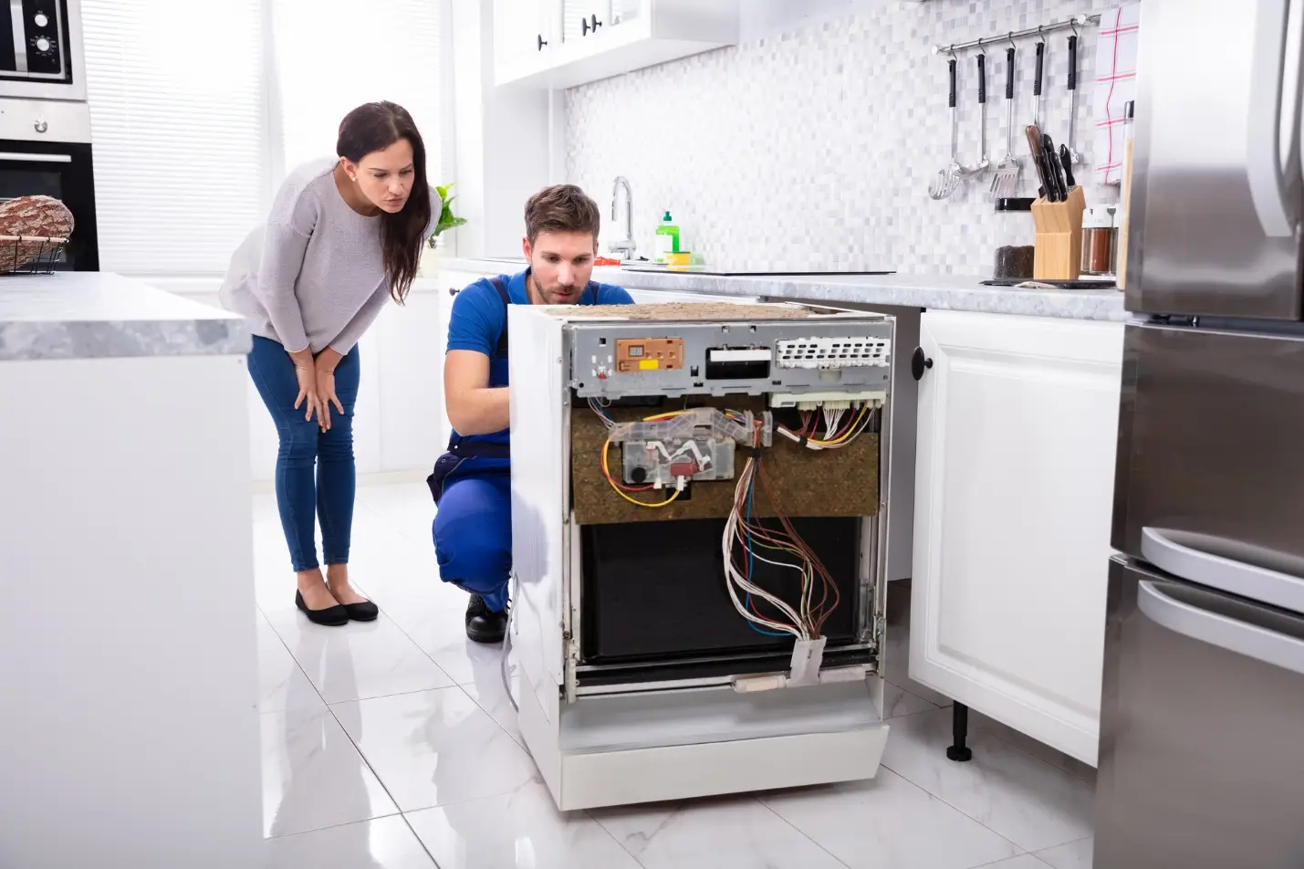 Man repairing a dishwasher with a woman observing in a modern kitchen setting