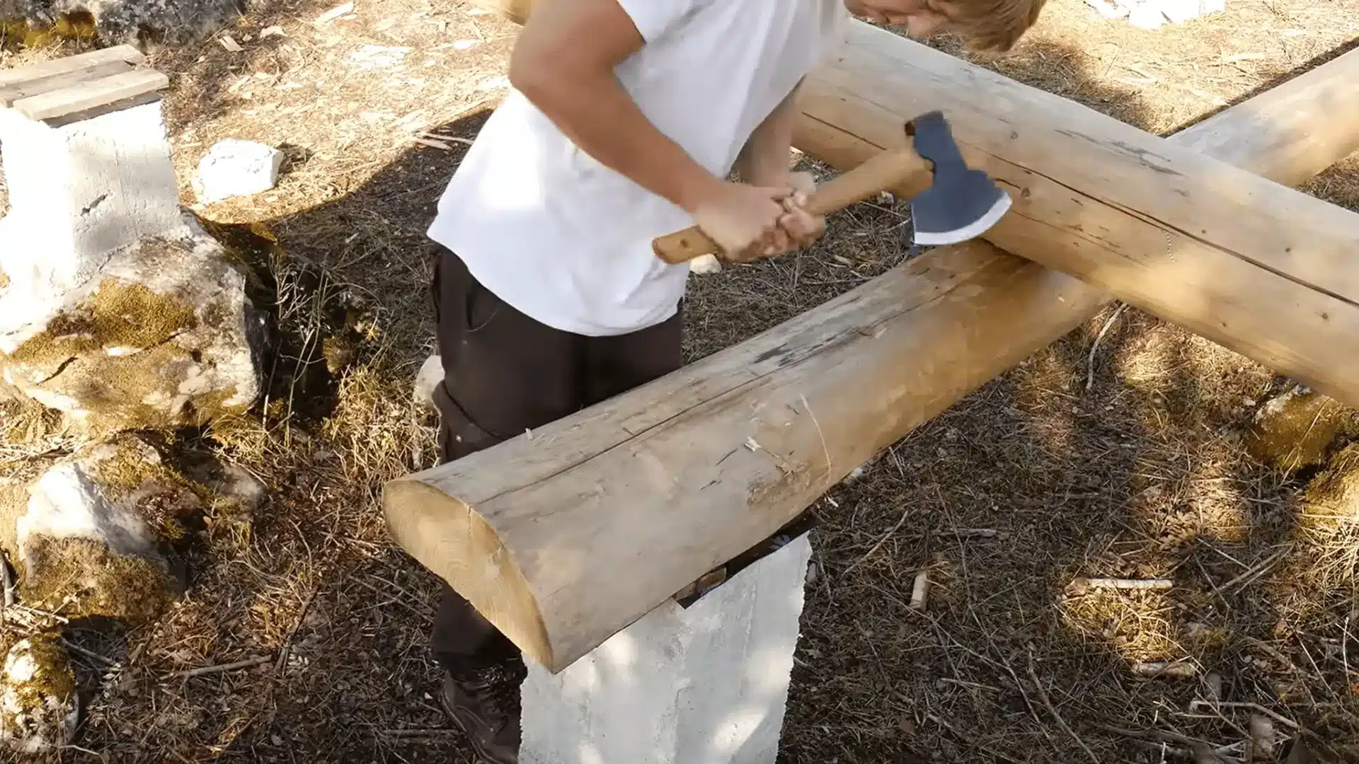 Shaping a foundation log with a broad axe where two logs meet above the concrete supports