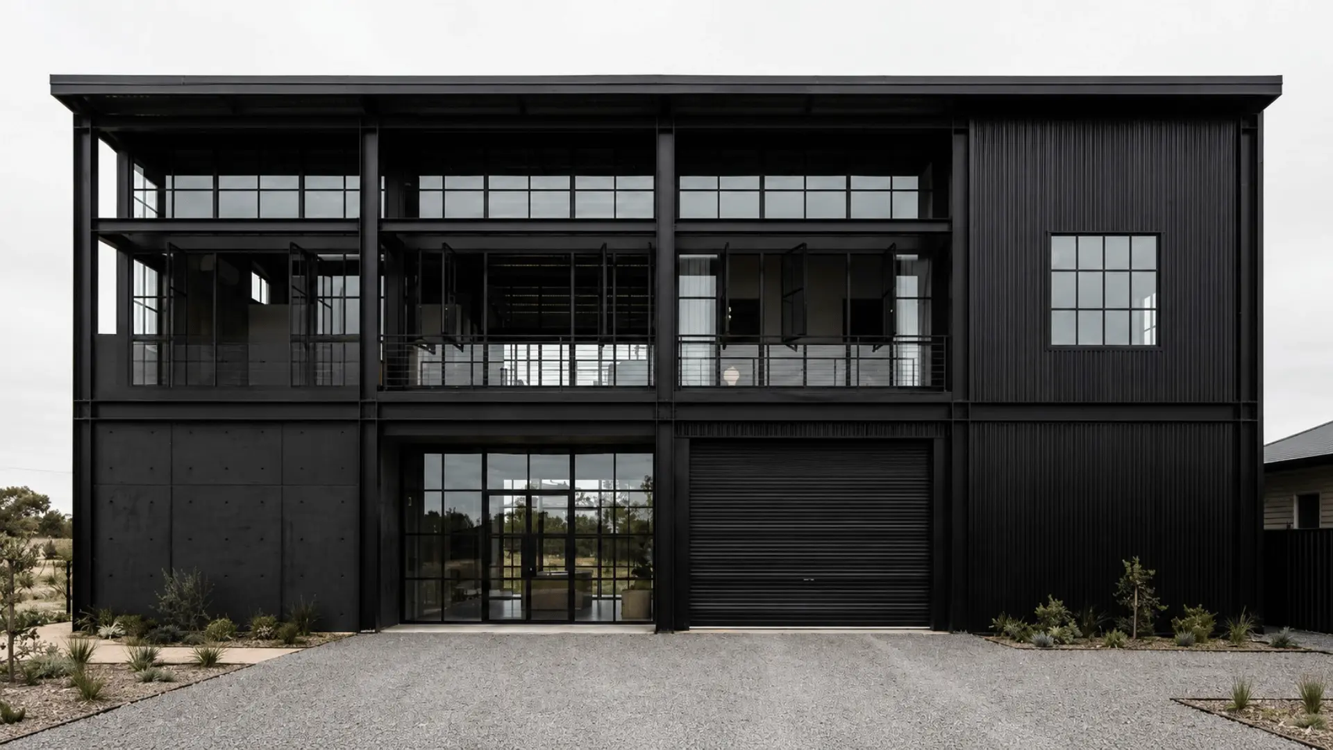 Full front view of a black industrial loft-style house with steel-framed glazing, exposed structure, and broad openings