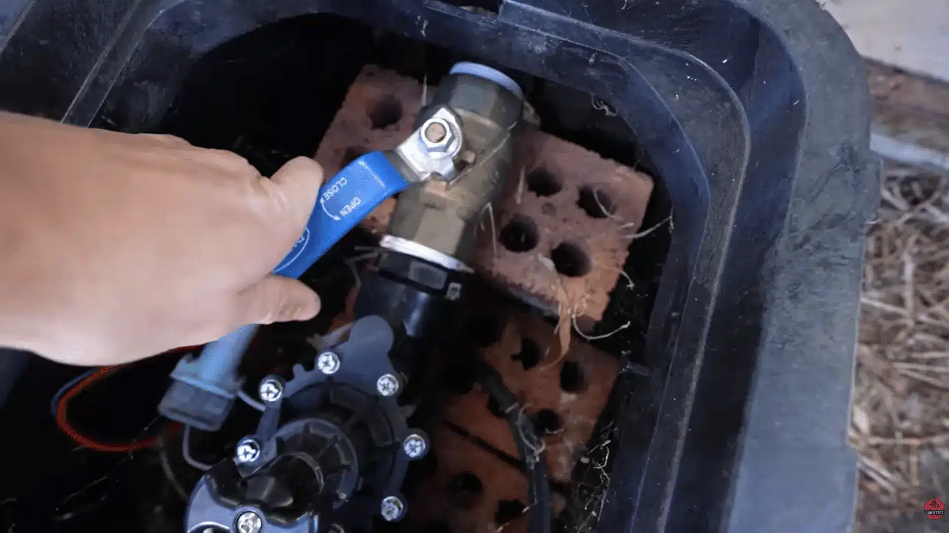 Hand turning a blue-handled shut-off valve inside an irrigation box before connecting the sprinkler main line.