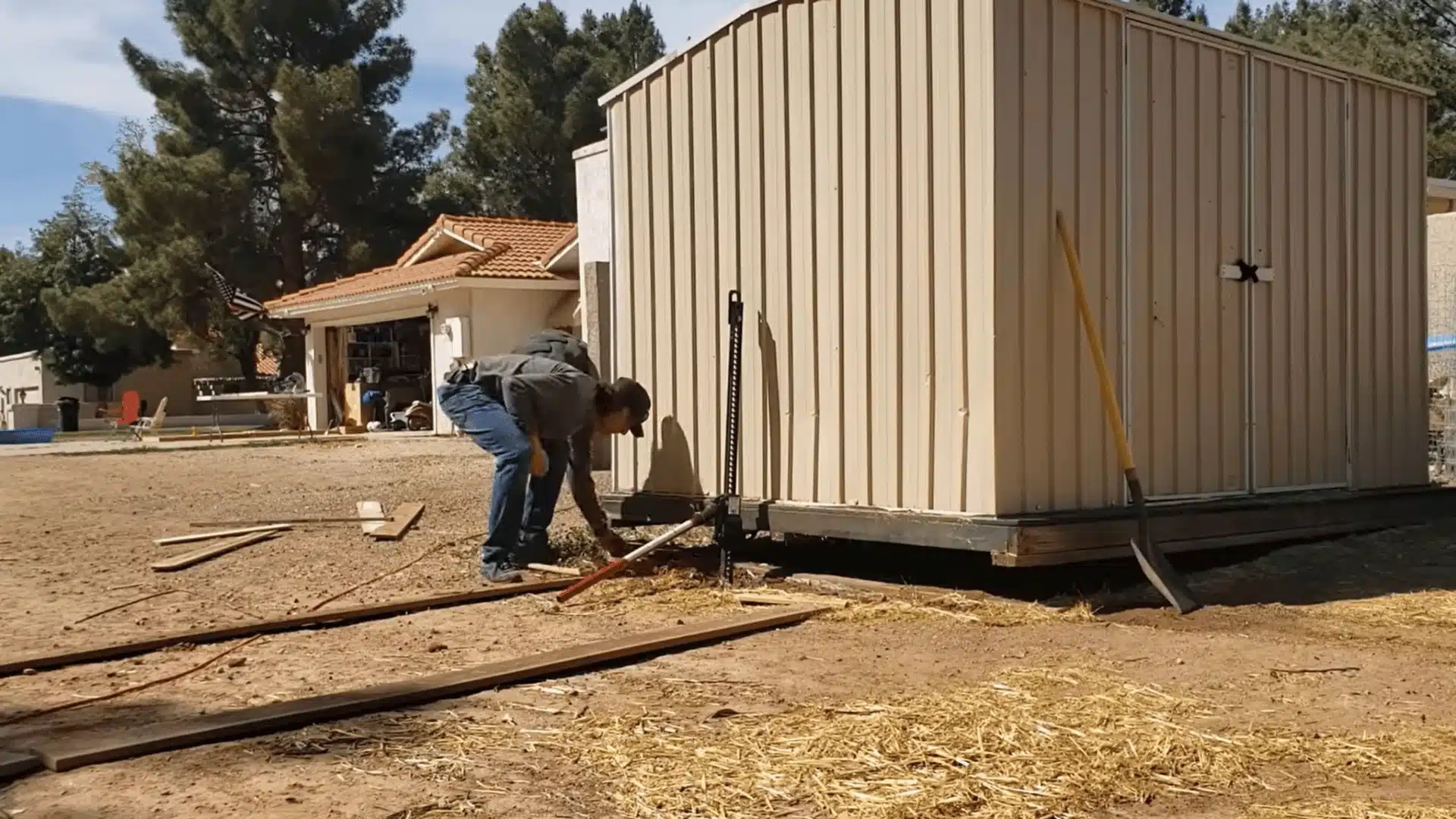 High-lift jack positioned under the edge of a metal shed to raise it before placing rollers underneath.