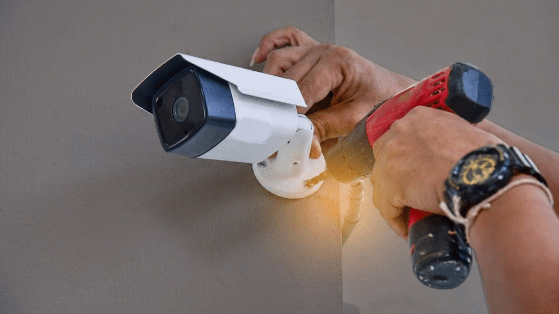 person uses a red power drill to mount a white security camera onto a dark gray wall