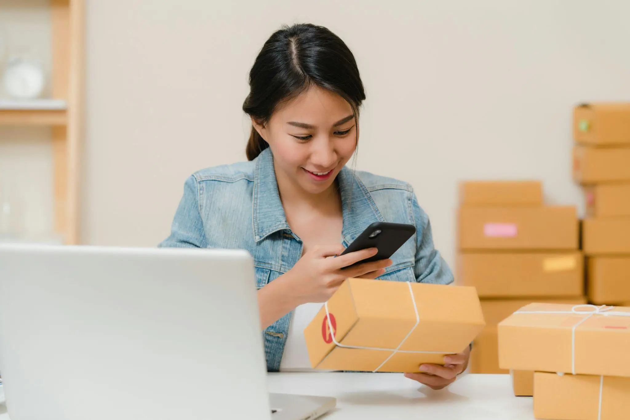 Woman in denim jacket using smartphone to photograph packages in home office setting