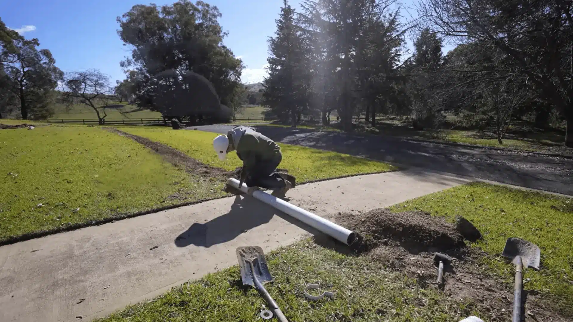 Installing a white PVC sleeve beneath a concrete path for sprinkler system irrigation pipes.