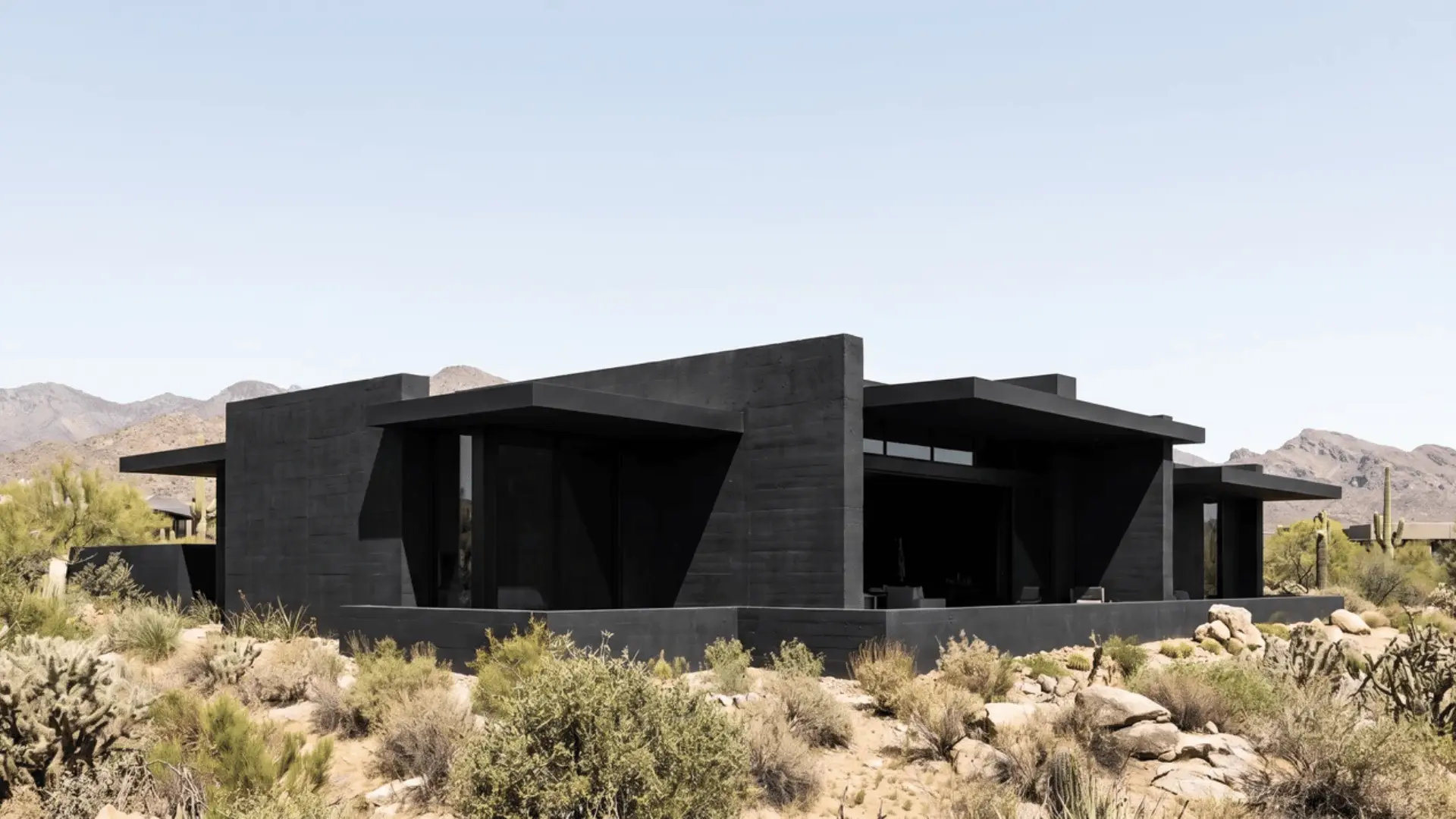 Low black desert house with broad rooflines, recessed openings, thick walls, and shaded glazing