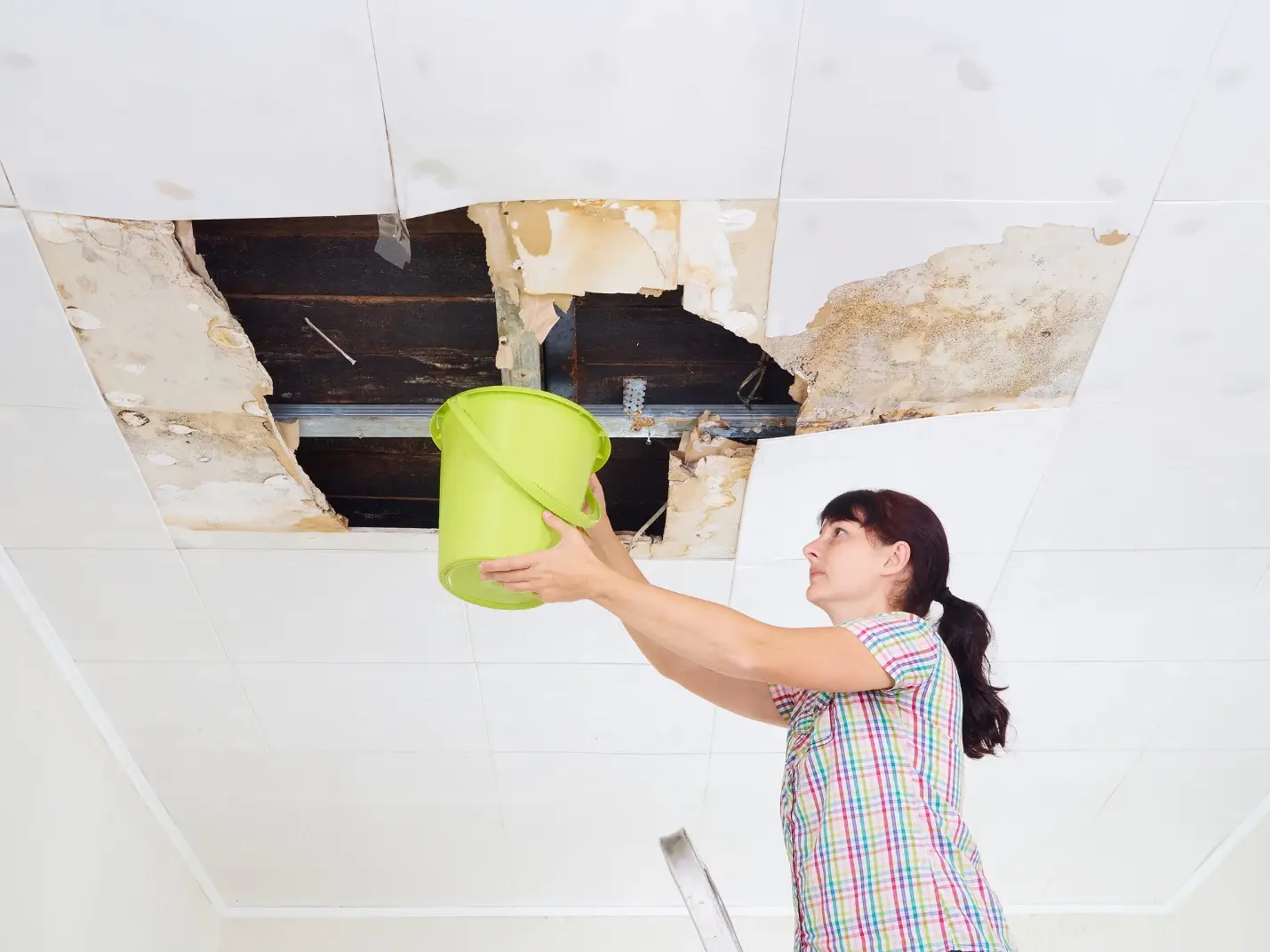 Woman holding green bucket under water-damaged ceiling indoors