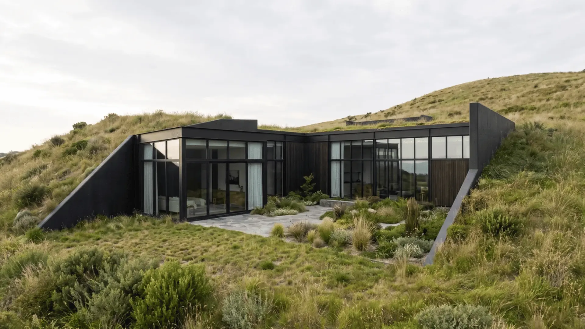 Partially buried black earth-sheltered house with exposed glazing, dark walls, and landscape covering part of the structure
