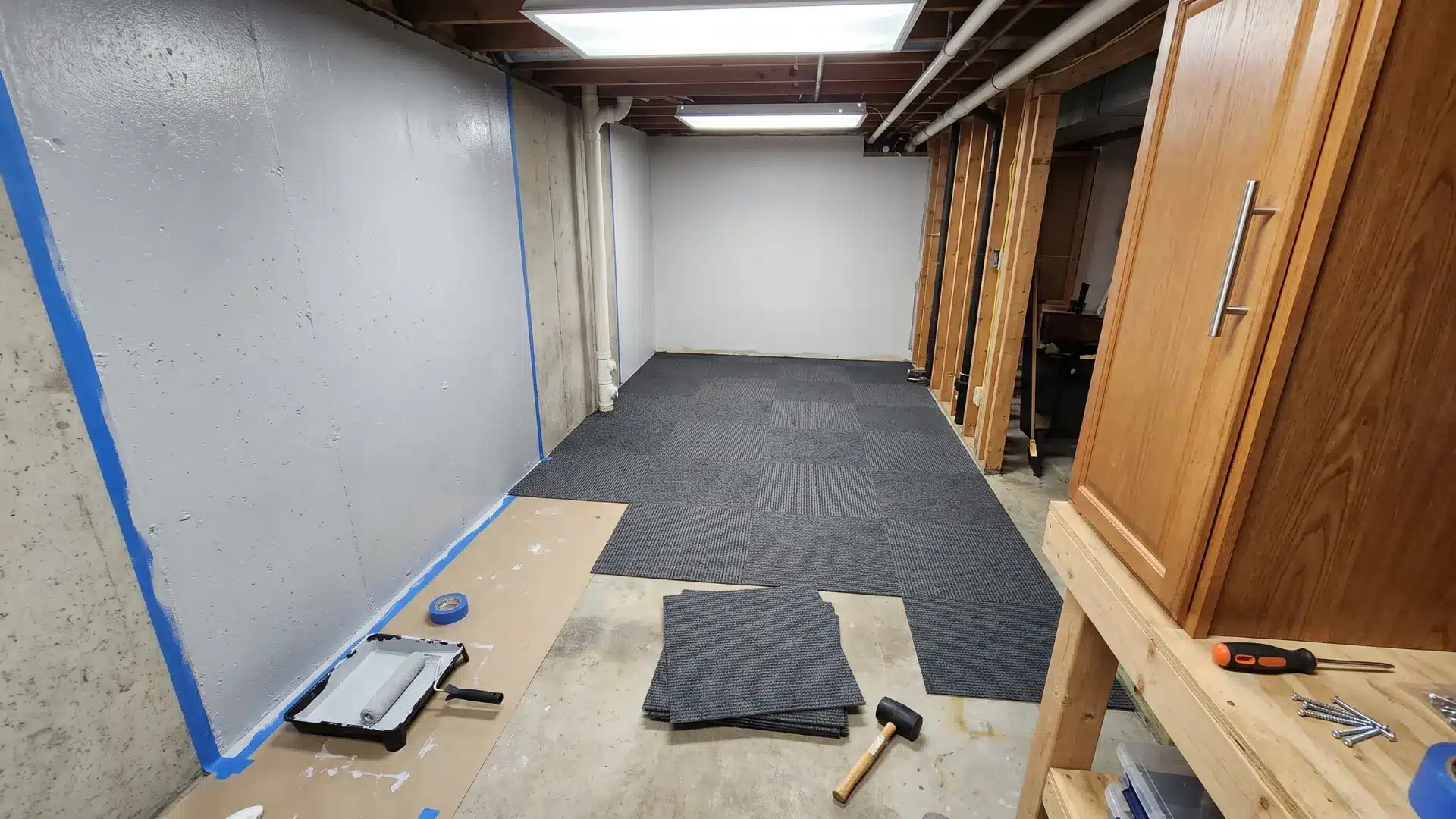 Small basement renovation with painted walls, carpet tiles, exposed ceiling pipes, and tools on the concrete floor.