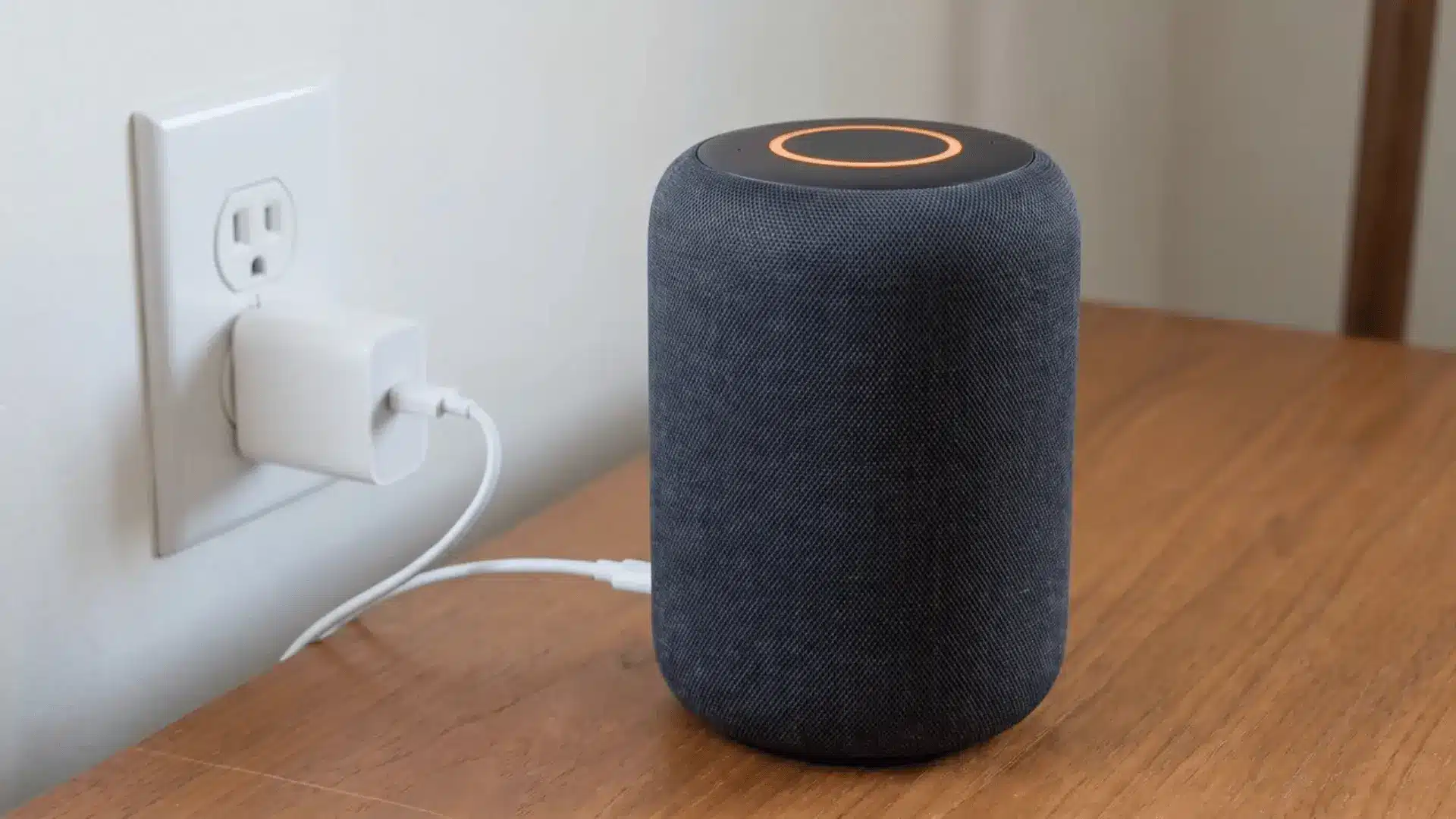Smart speaker plugged into a wall outlet with its power adapter and a solid orange light ring