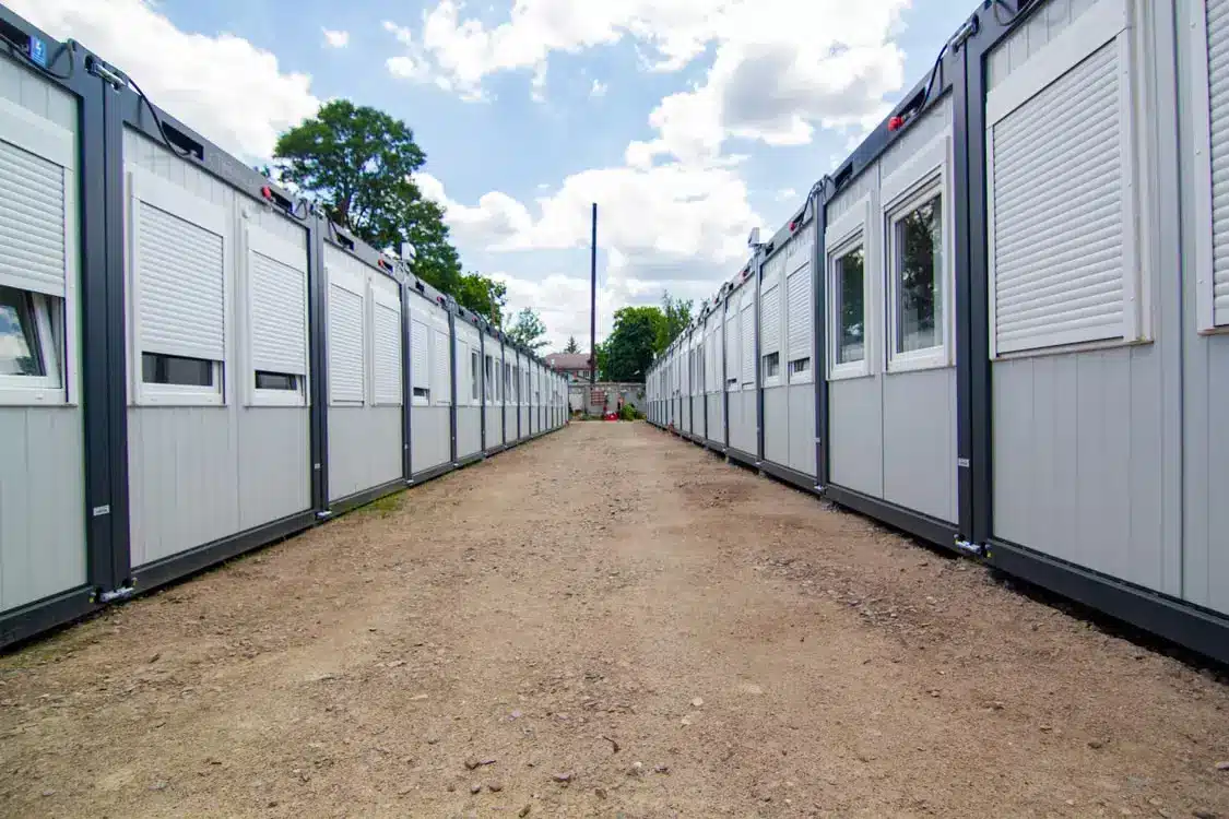 Row of gray modular buildings with closed shutters on dirt path under cloudy blue sky