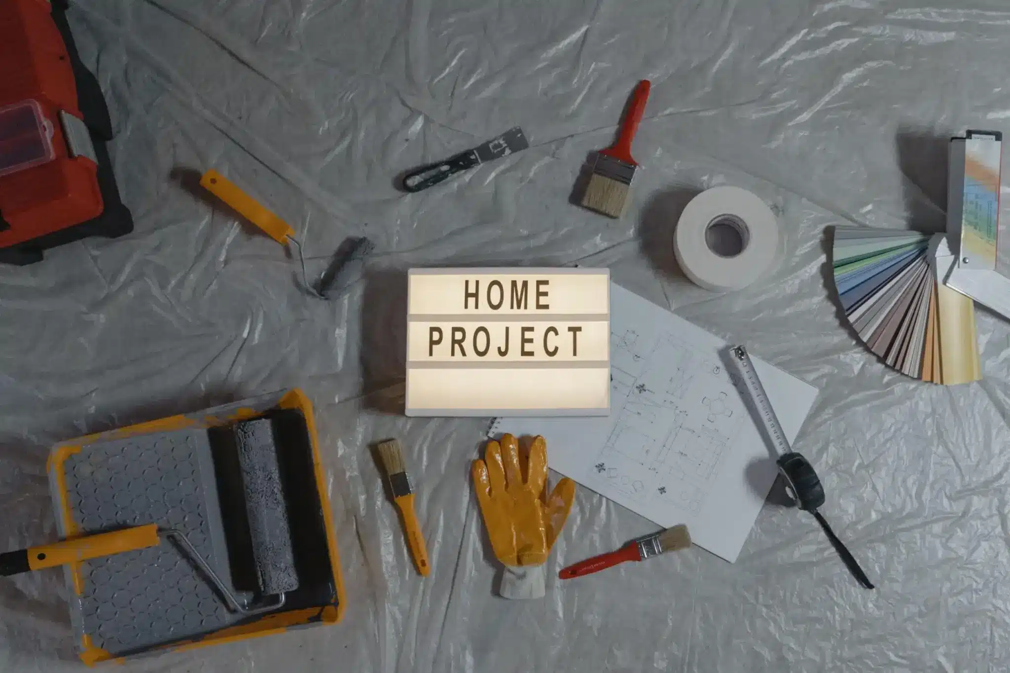 Lightbox with "Home Project" surrounded by paint tools and color samples on plastic-covered floor
