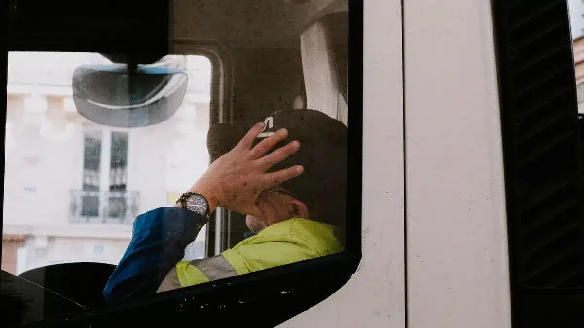 Person wearing a cap and reflective jacket inside a truck cab, hand on head