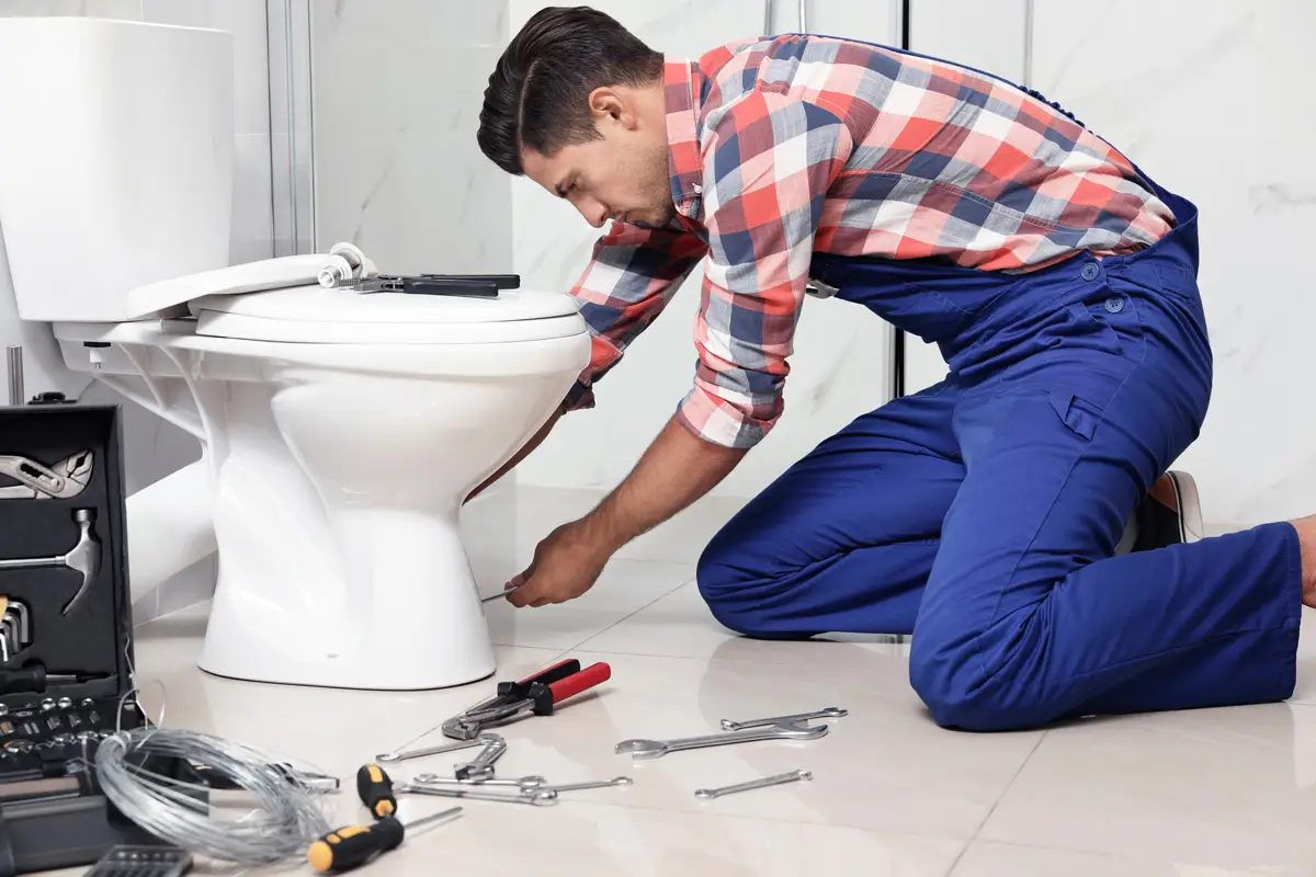 Plumber in blue overalls fixing toilet with tools scattered in modern bathroom