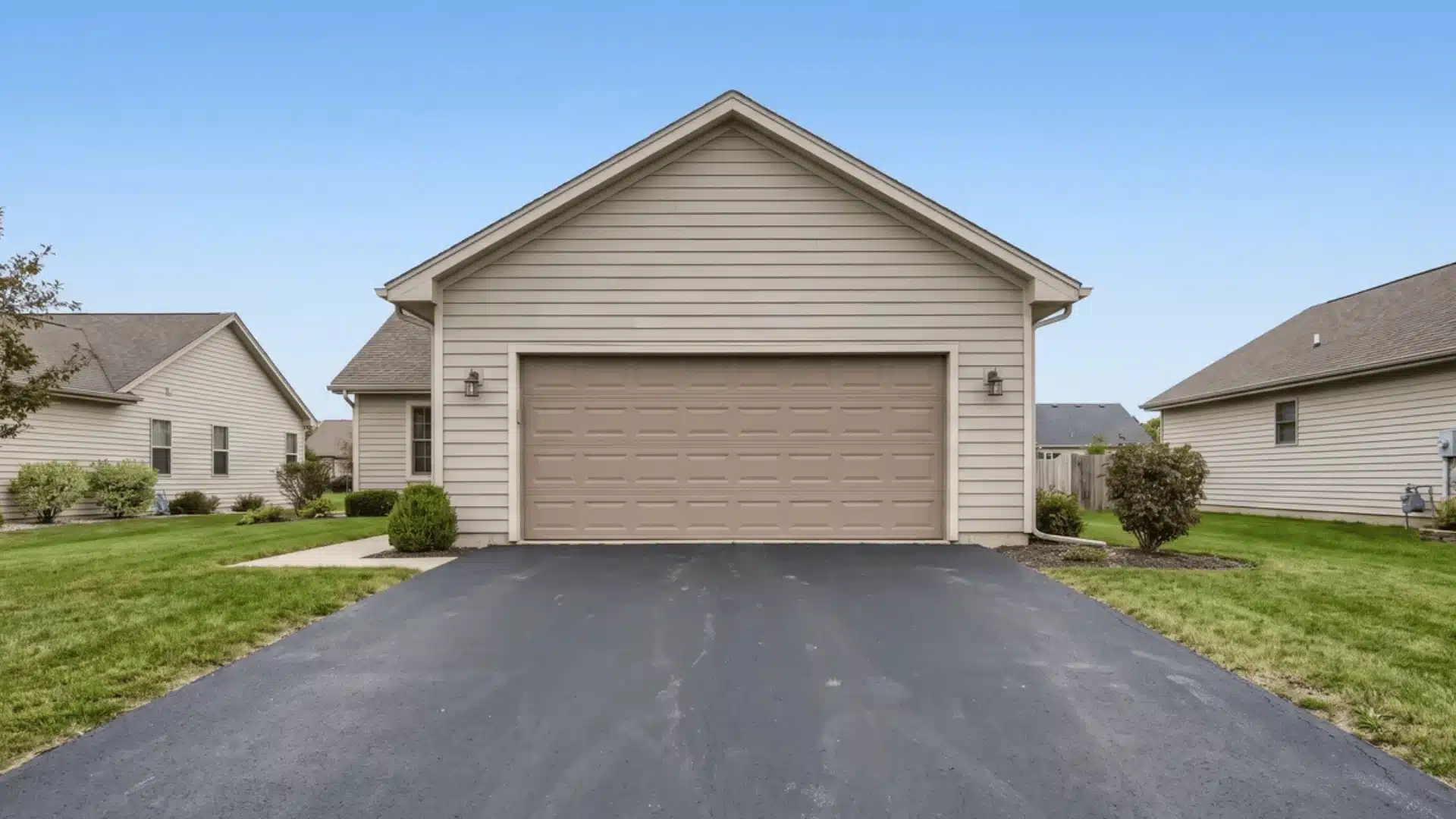 Detached two-car garage with beige siding, closed overhead door, paved driveway, green lawns, and neighboring suburban homes under blue sky