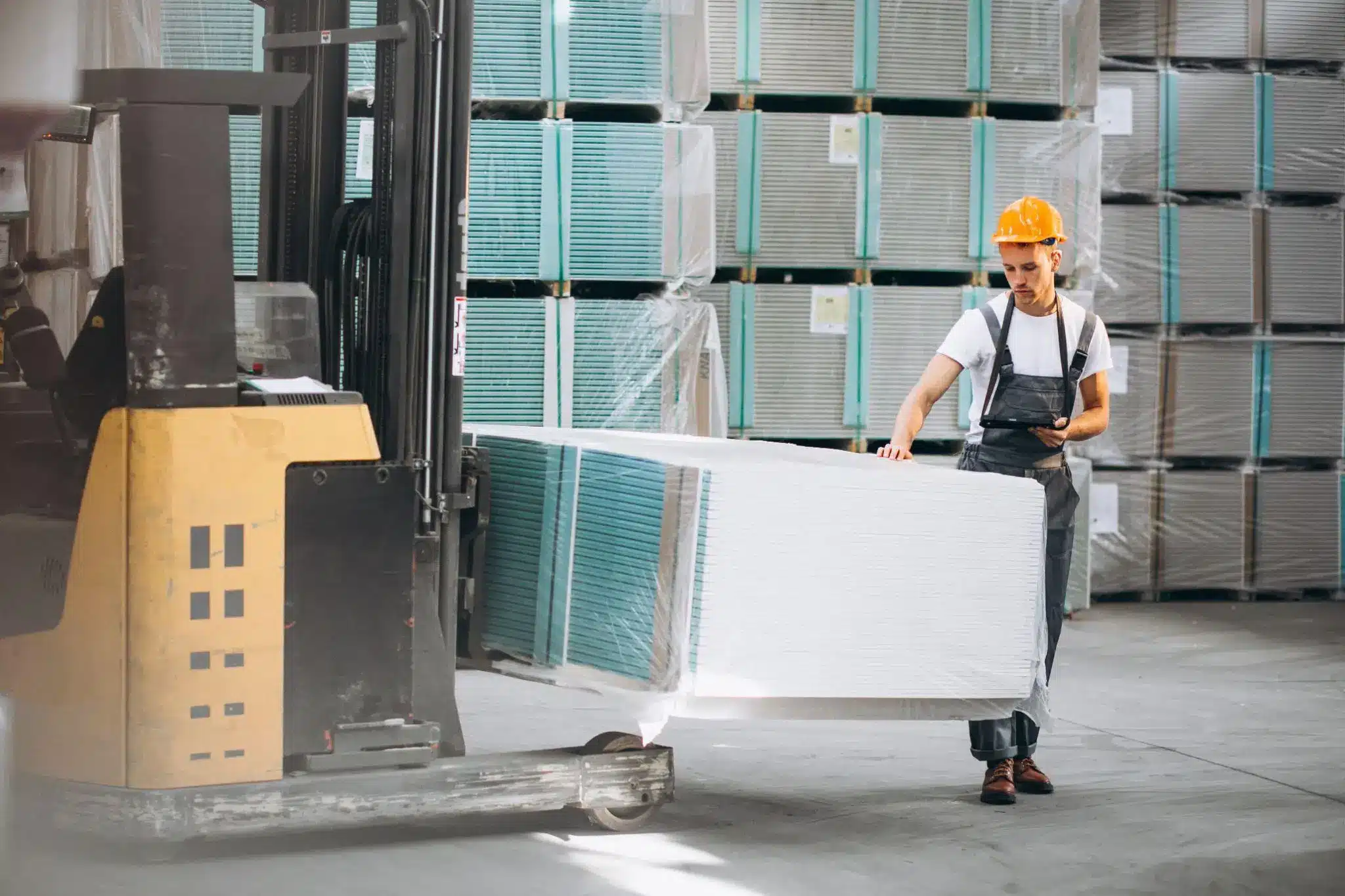 Worker in orange hard hat operating forklift in warehouse with stacked drywall panels