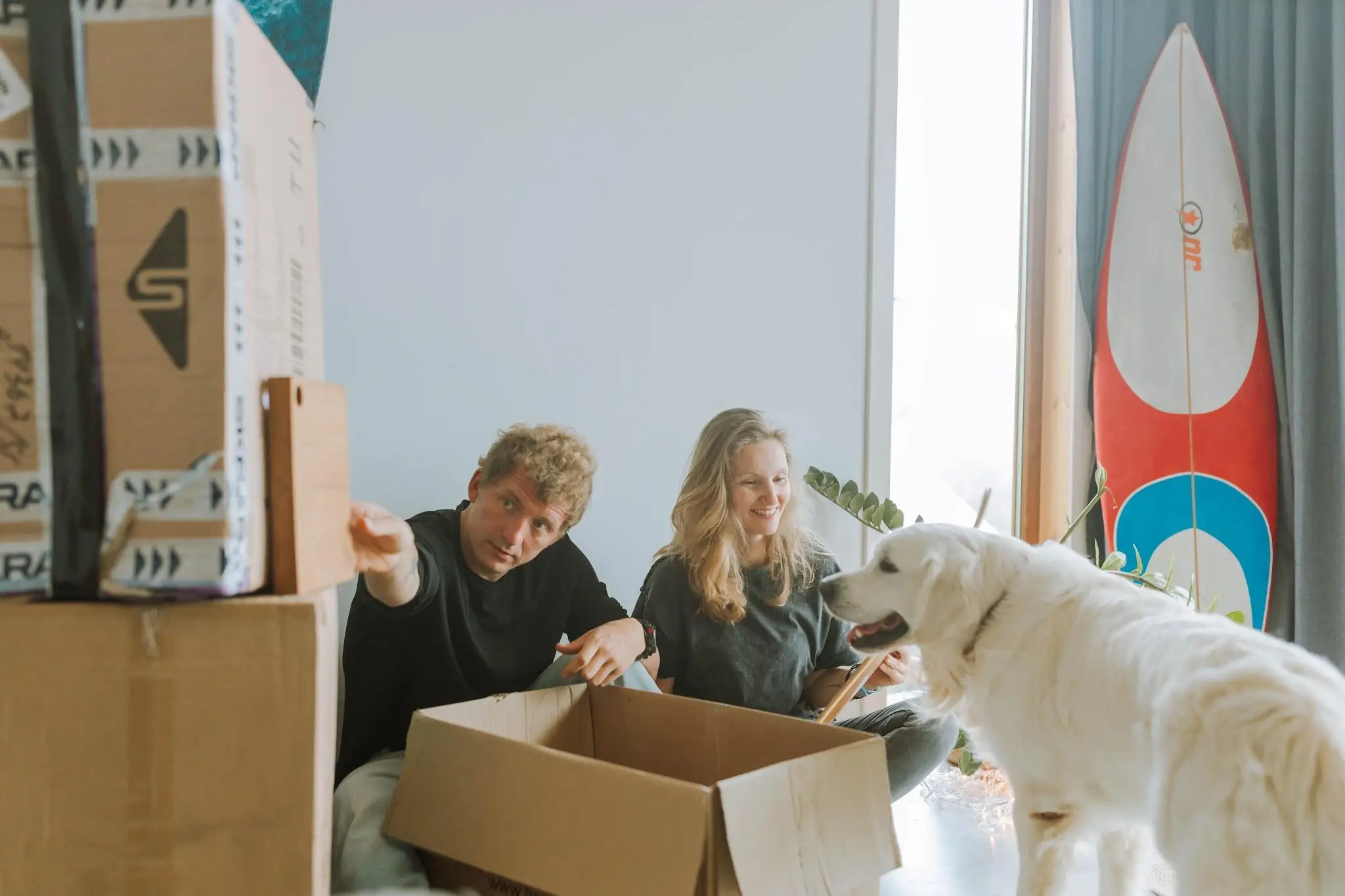 Couple unpacking boxes with a dog in a bright living room, surfboard leaning against wall