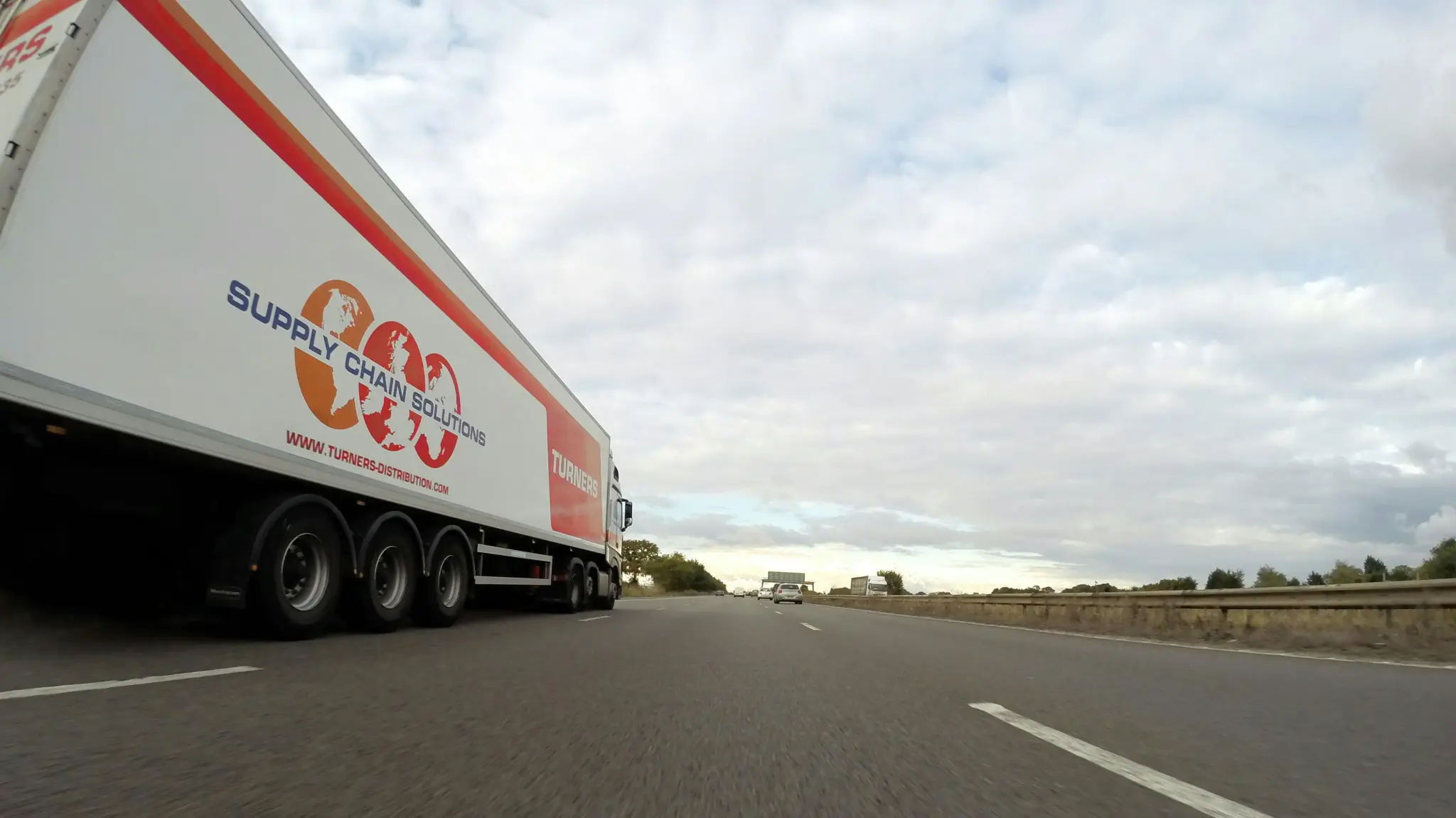 Truck with supply chain logo driving on highway under cloudy sky
