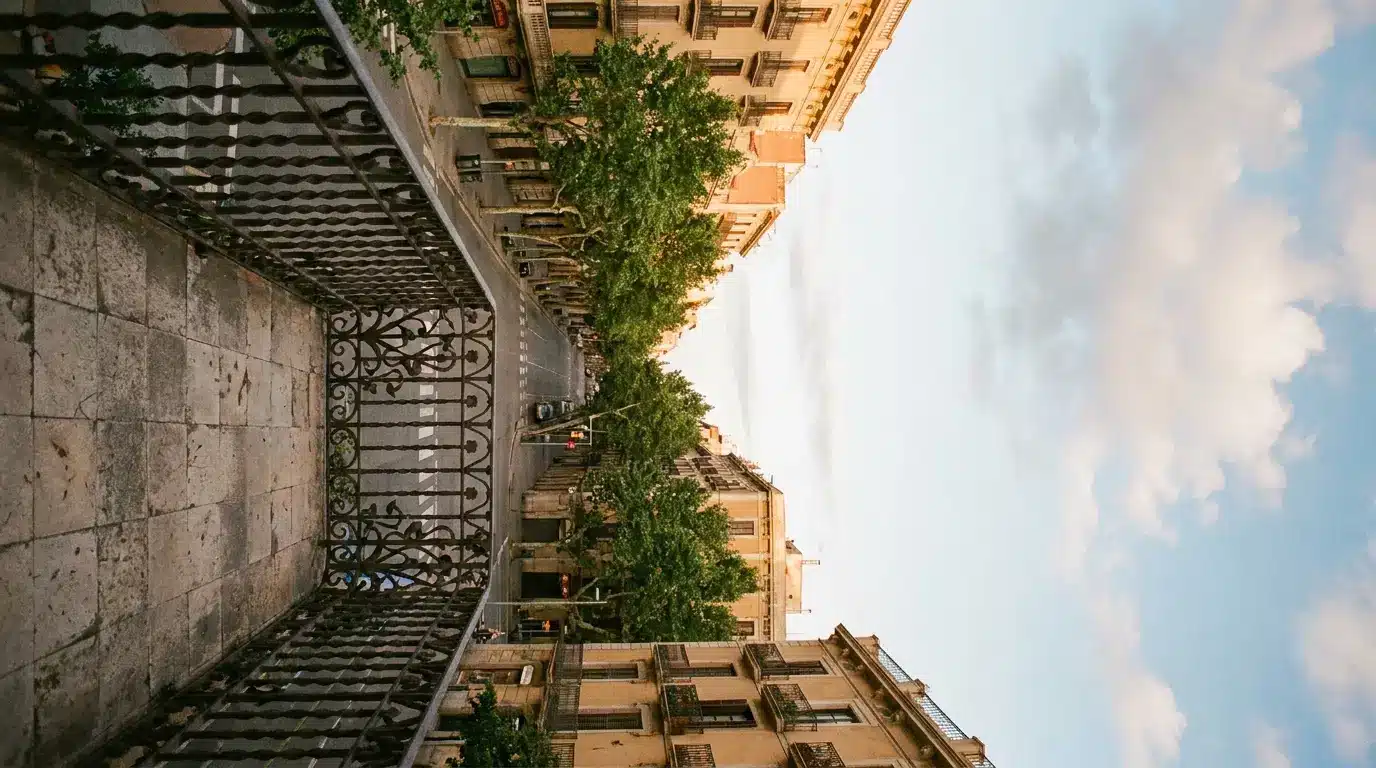 Urban street view from balcony with ornate railing, lined with trees and buildings under blue sky