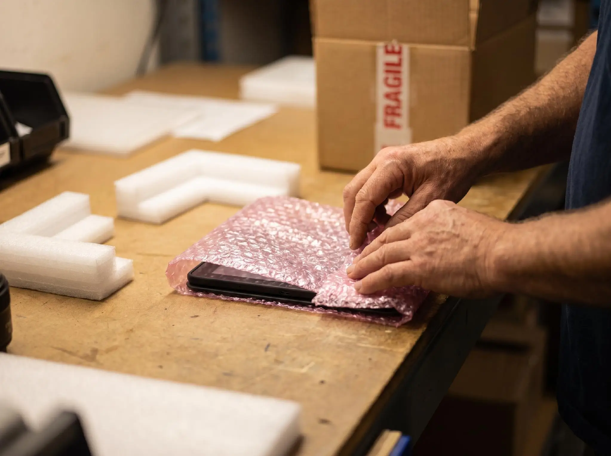 Hands wrapping a smartphone in pink bubble wrap on a wooden table in a workshop setting