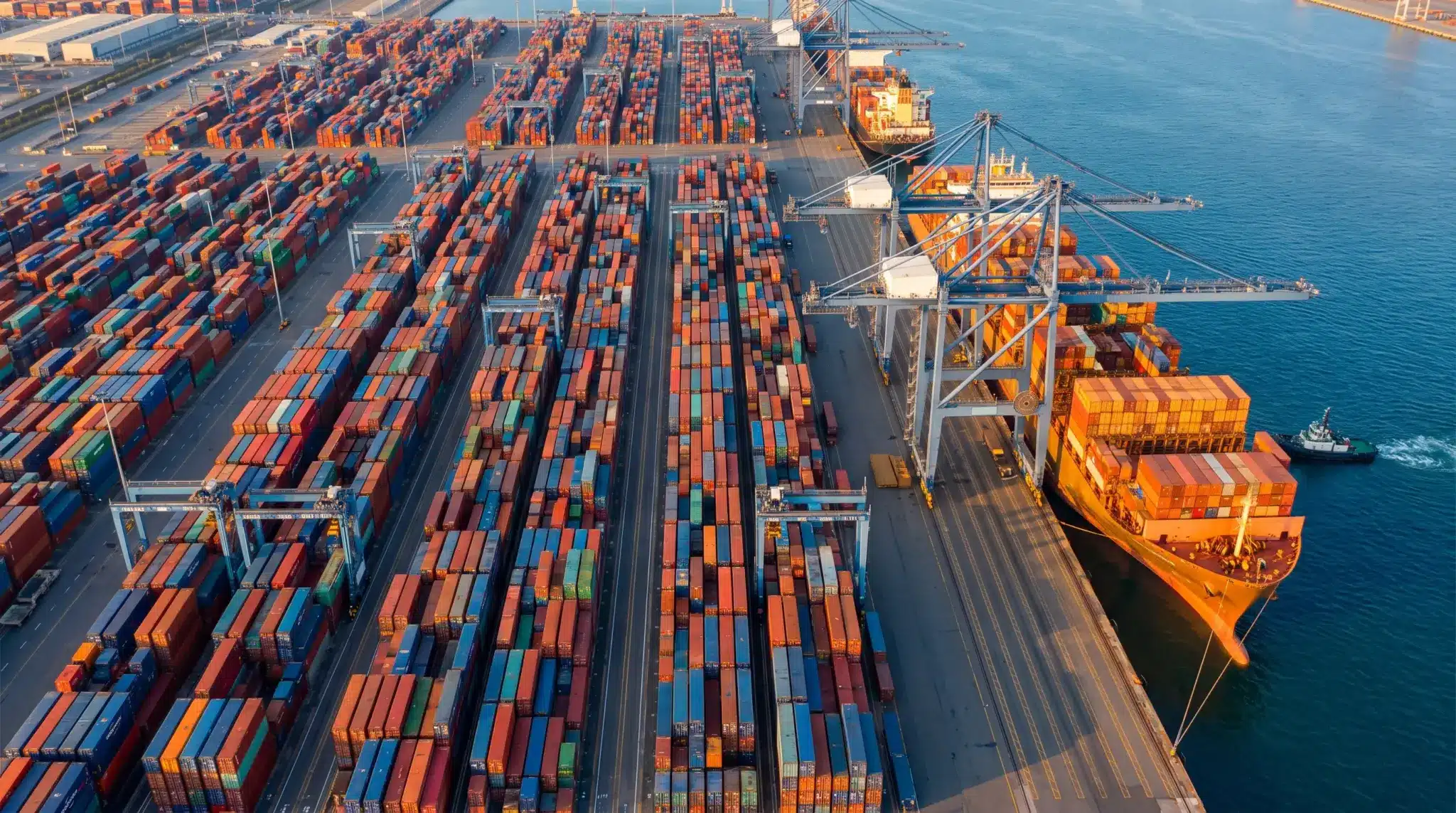 Cargo containers stacked at busy port terminal with cranes and cargo ship docked in bright sunlight