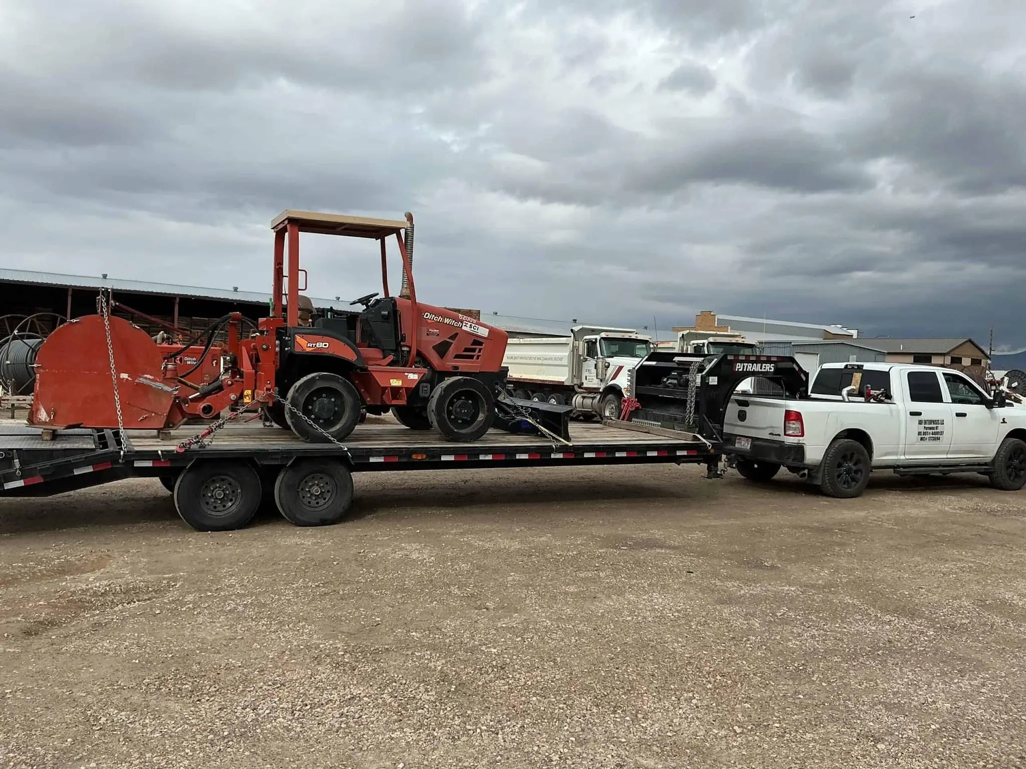 Red trencher on flatbed trailer hitched to white pickup truck under cloudy sky