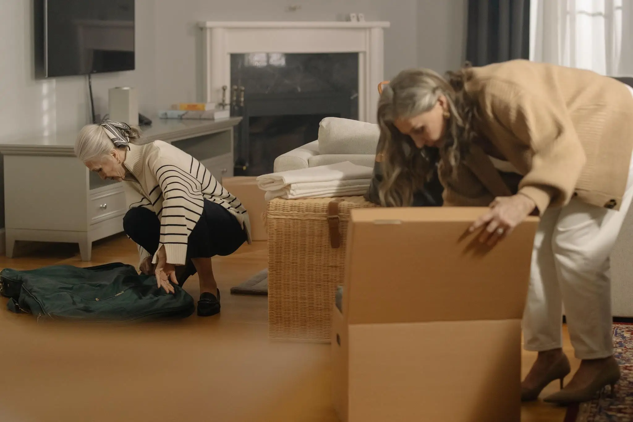 Two women packing items in cardboard box and suitcase in cozy living room
