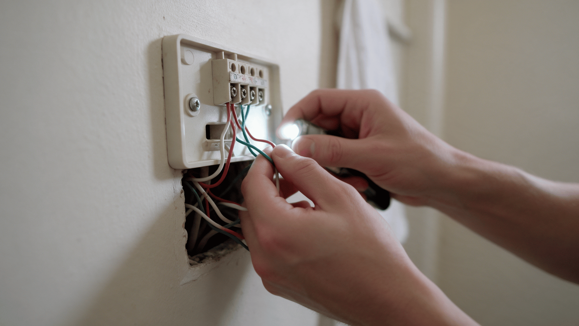 hands inspecting thermostat wiring behind a removed wall plate