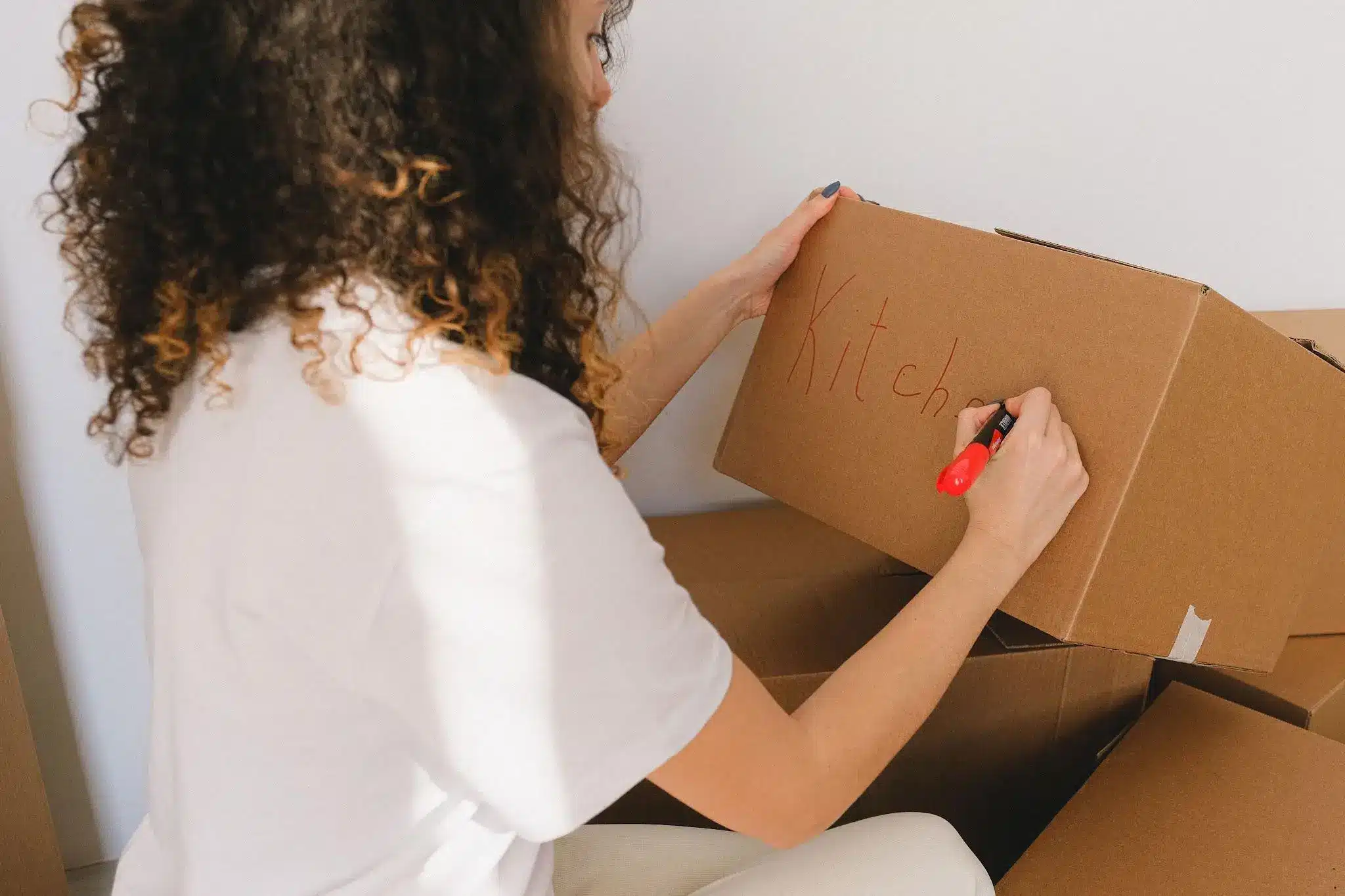 Woman writing "kitchen" on moving box with red marker in a bright room