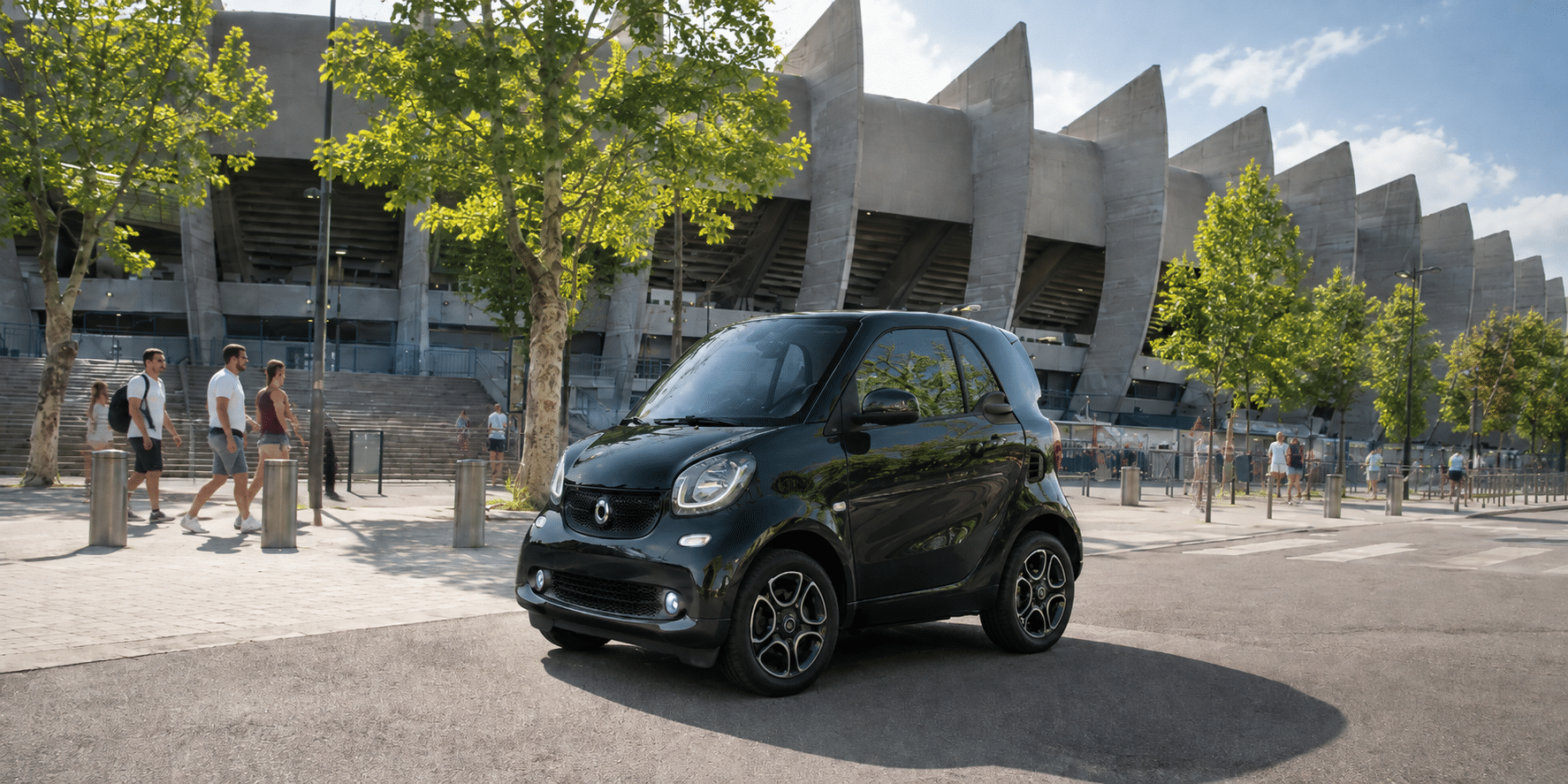 Black compact car parked outside modern stadium with trees and people walking nearby