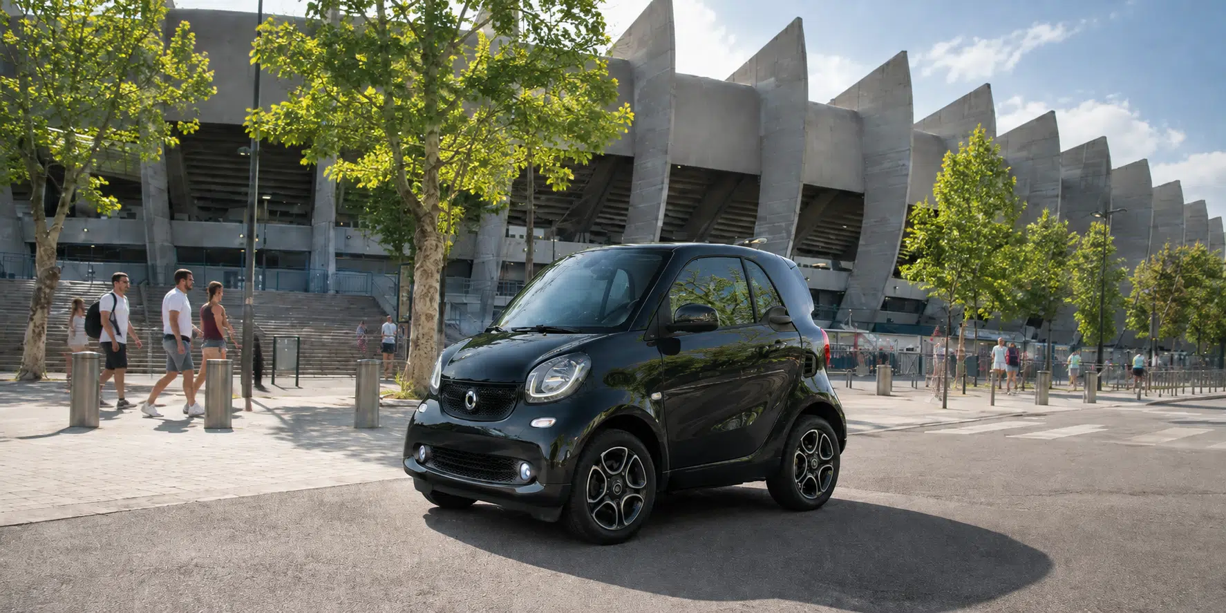 Black compact car parked outside modern stadium with trees and people walking nearby