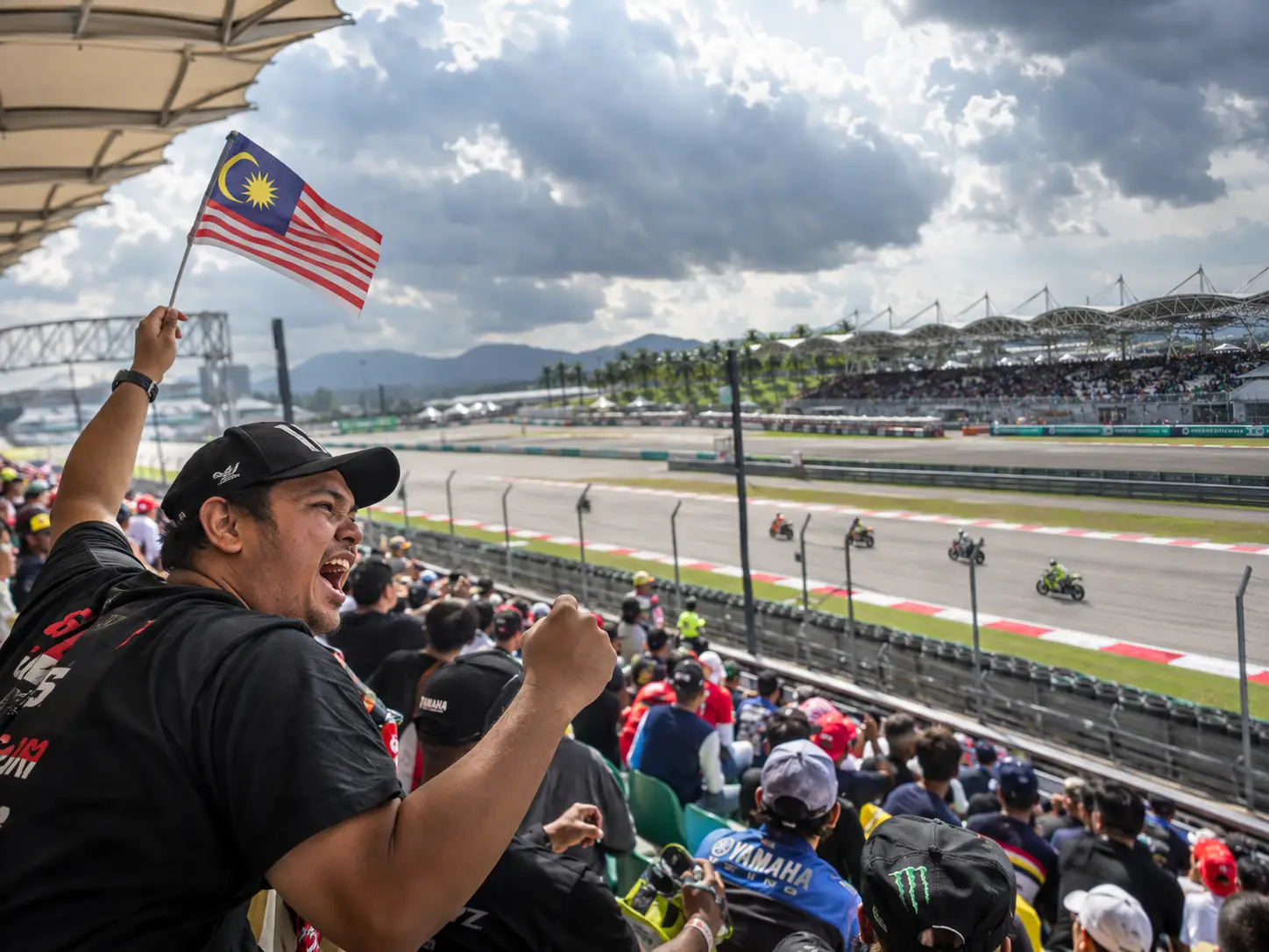 Race fan waving Malaysian flag at motorcycle race on sunny day in crowded grandstand