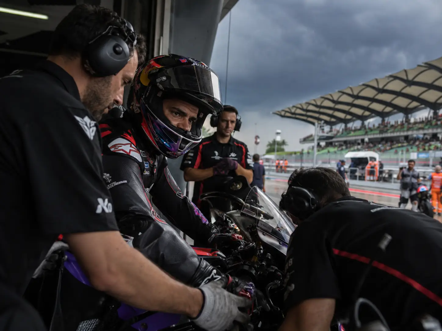 Motorcycle racer in helmet surrounded by mechanics in pit lane with cloudy sky overhead