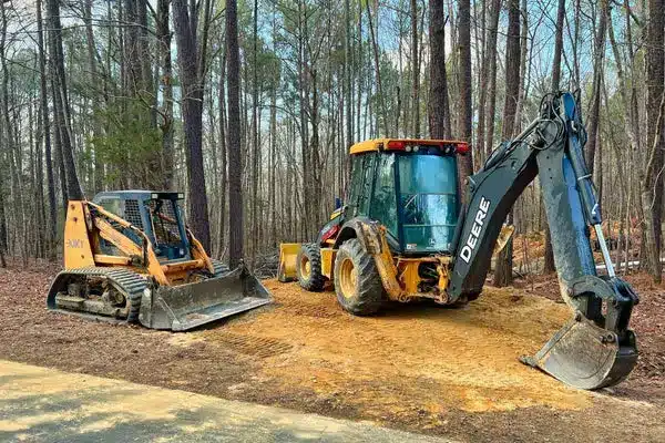 Two construction vehicles parked on a dirt path in a wooded area