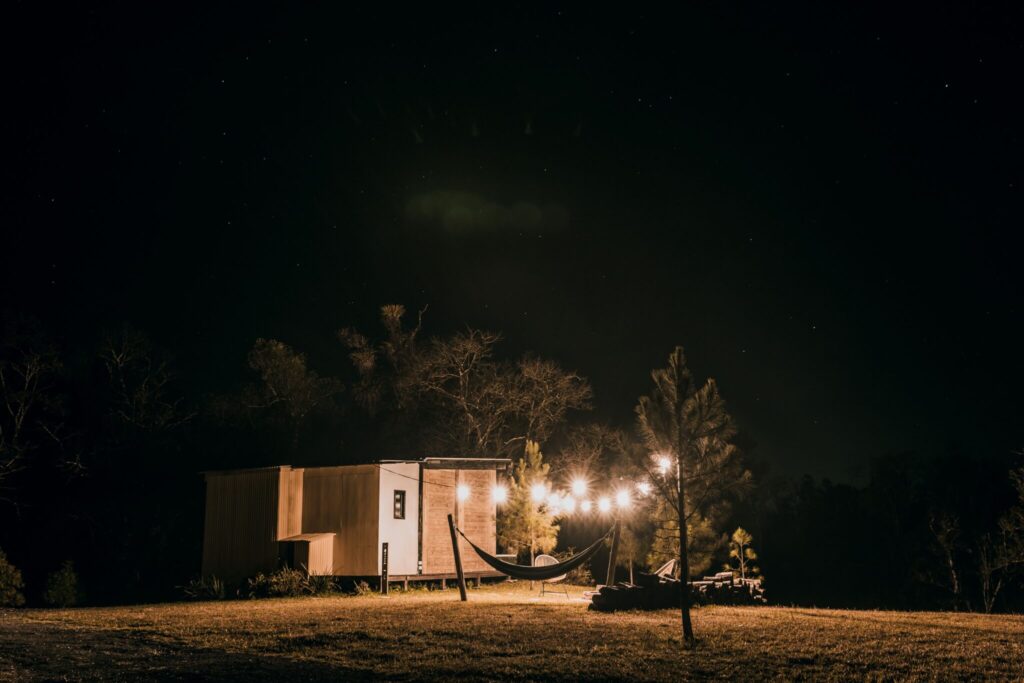 Hammock and lit tiny house on grassy field under starry night sky