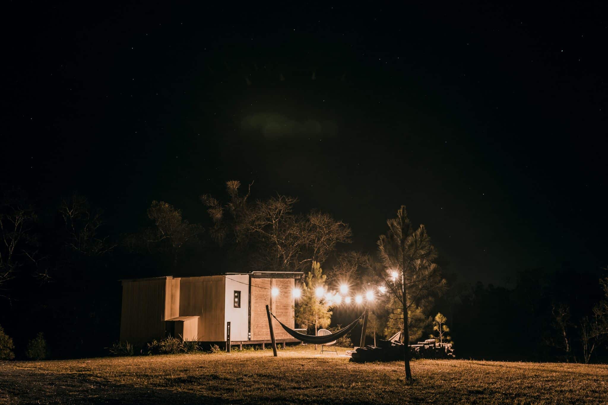 Hammock and lit tiny house on grassy field under starry night sky