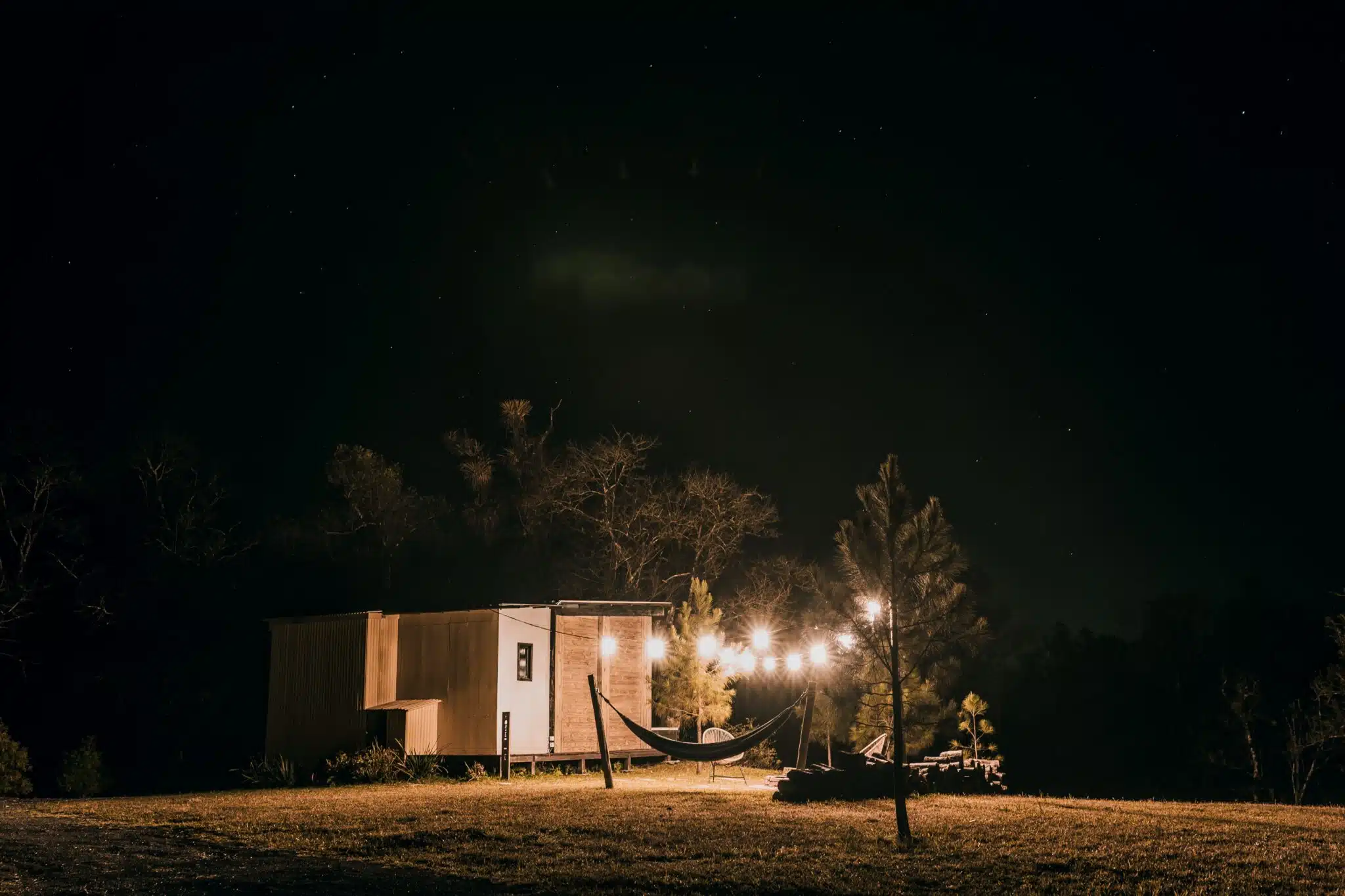 Hammock and lit tiny house on grassy field under starry night sky