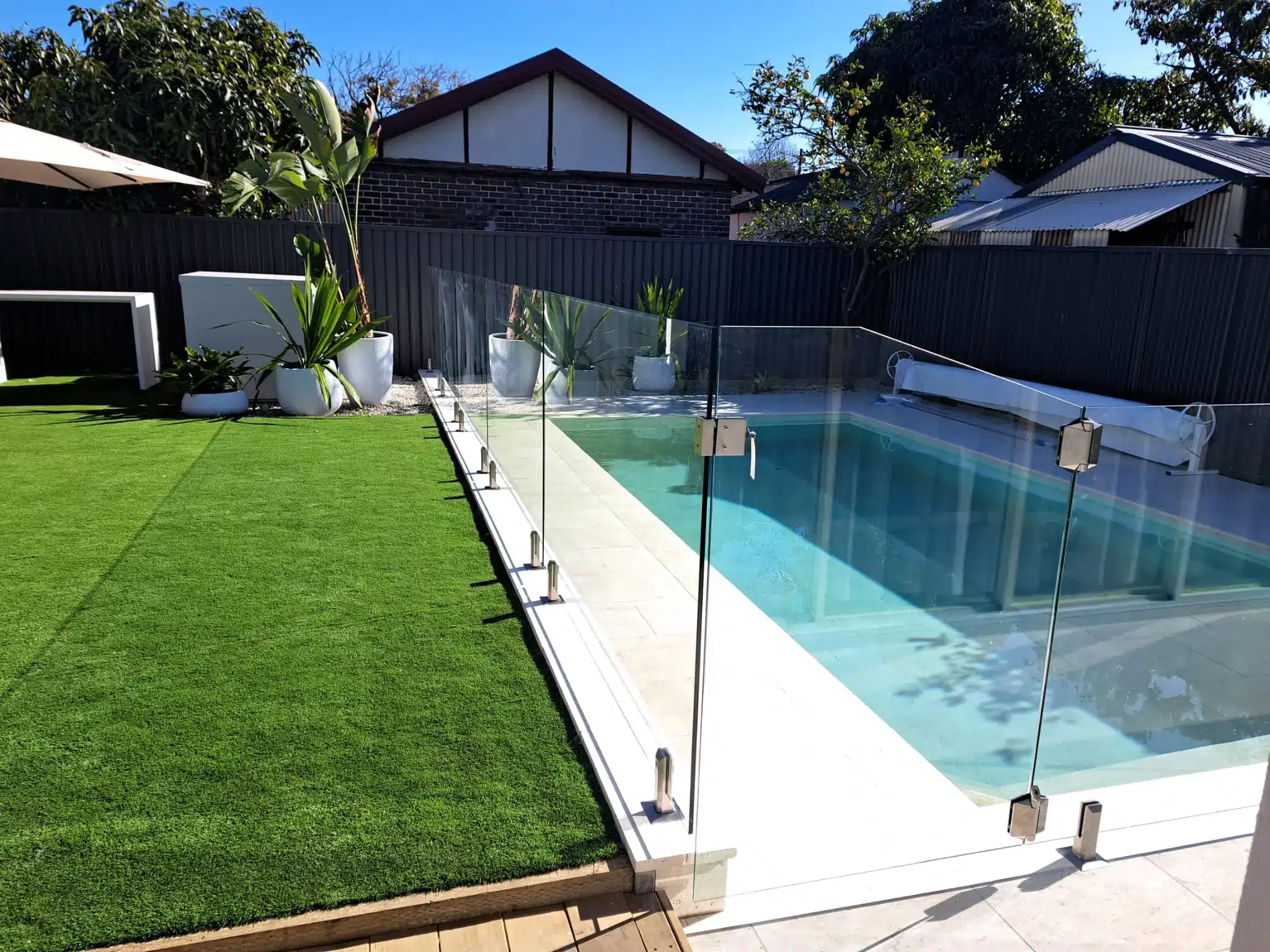 Backyard pool with glass fencing and artificial grass lawn under clear blue sky