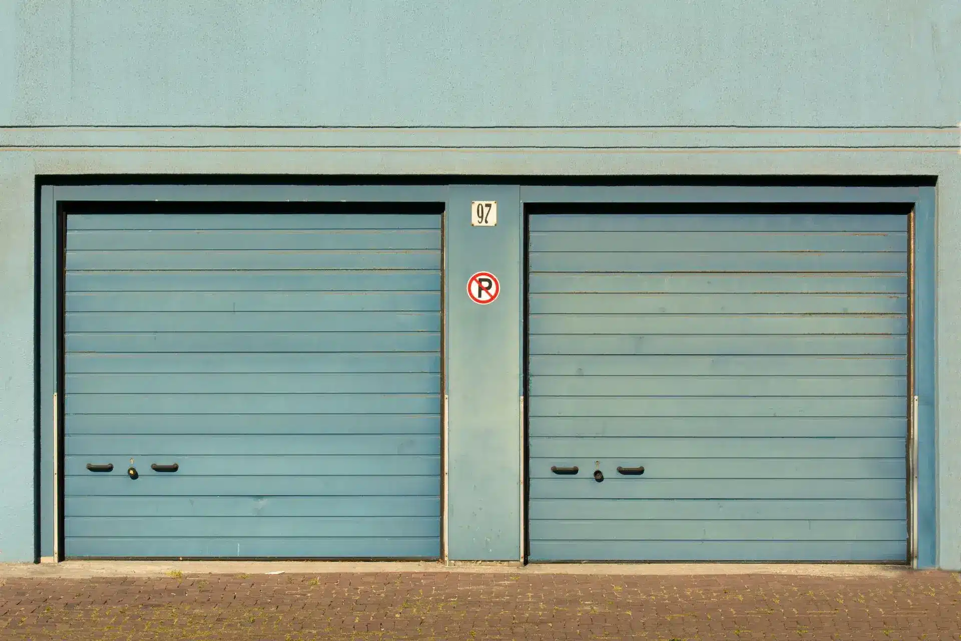 Two closed blue garage doors with no parking sign on brick driveway